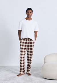 Weißes T-Shirt mit kurzen Ärmeln, kombiniert mit braunen und cremefarbenen karierten Schlafanzughosen mit elastischem Bund und Kordelzug. Der Boden ist hellgrau.