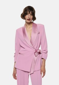Lyserød skræddersyet blazer med satin revers og bælte detalje, der har lange ærmer og lommer. Glat tekstur, struktureret design og tætsiddende silhuet.