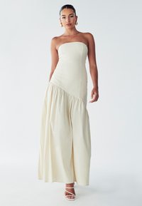 Robe maxi beige sans bretelles avec un corsage ajusté et un ourlet asymétrique froncé. Texture lisse avec un tissu fluide, associé à des talons blancs à lanières.