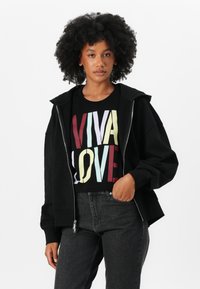 Čierna mikina na zips s veľkými prednými vreckami, pod ktorou je čierne tričko s grafikou, na ktorom sú farebné nápisy "VIVA LOVE". Tmavé džíny.