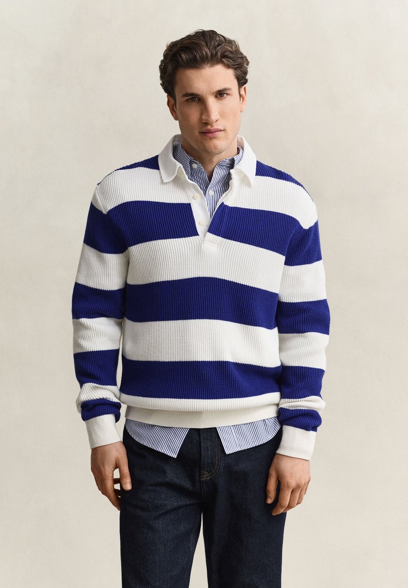 Jeune homme portant un pull rayé horizontal bleu et blanc par-dessus une chemise à rayures bleues avec boutons et un jean foncé sur un fond uni.