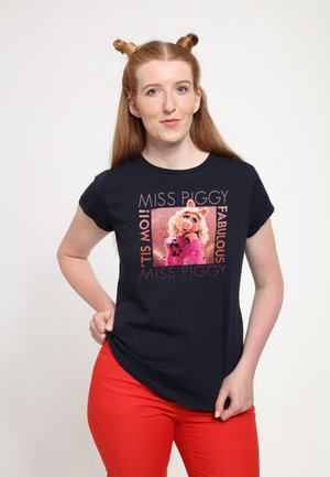 Granatowa koszulka z grafiką Miss Piggy w różowym płaszczu, otoczona napisem "MISS PIGGY FABULOUS! 'TIS MOI!" Noszone czerwone spodnie.