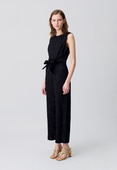 Jumpsuit nero senza maniche con un pattern floreale testurizzato, cintrato in vita, abbinato a sandali con tacco dorati con cinturini.