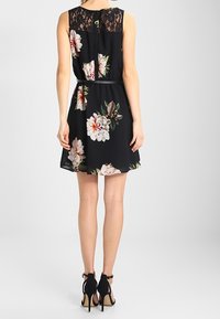 Černé šaty s květinovým vzorem, krajkovým zadním dílem, bez rukávů, s úzkým pasem a rozšířenou sukní, s mírným leskem a detailem otevřeného zadního dílu.