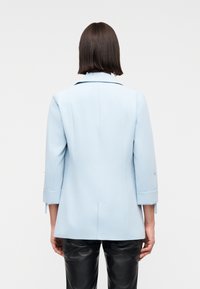 Άτομο με κοντά σκούρα μαλλιά, που φοράει γαλάζιο σακάκι με μανίκια 3/4 και μαύρο δερμάτινο παντελόνι, γυρισμένο πλάτη σε λευκό φόντο.