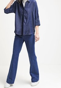 Camicia di denim blu scuro con vestibilità rilassata, colletto button-down e maniche arrotolate; abbinata a jeans larghi blu scuro e sneakers bianche.