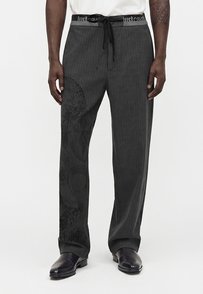 Homme portant un pantalon gris foncé à fines rayures avec un motif subtil de crâne, des chaussures en cuir noir et une chemise blanche, debout devant un fond uni.