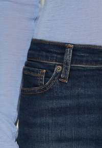 Mörkblå denimjeans med orange sömnad, med en ficka fram och knappstängning; kombinerat med en ljusblå långärmad skjorta.