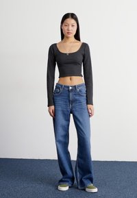 Crni rebrasti crop top dugih rukava u kombinaciji s ležernim plavim trapericama. Model nosi tenisice s akcentima u zelenoj boji, uz neutralnu pozadinu.