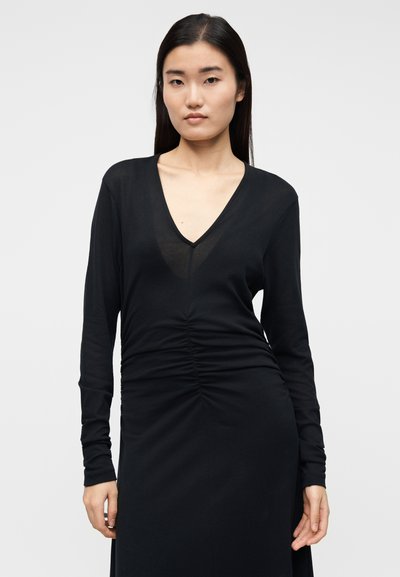 Μαύρο φόρεμα με μακριά μανίκια, βαθύ V-λαιμόκοψη και πιέτες κατά μήκος του εμπρόσθιου μέρους. Κατασκευασμένο από απαλό, ελαστικό ύφασμα με λεία υφή.