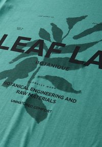 Groen textiel met donkere bladafdrukken en zwarte tekst met de woorden "Leaf Supply Co," "Botanique," en "Botanical engineering and raw materials."