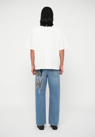 Persona con cabello oscuro hasta los hombros, viste una camiseta blanca de gran tamaño y unos pantalones vaqueros anchos azules con un gran parche floral oscuro en la pierna izquierda trasera.