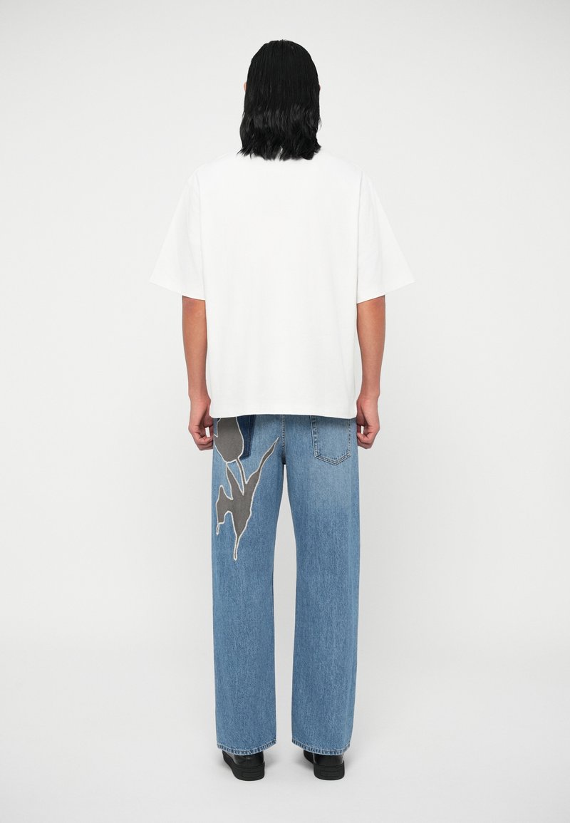 Eine Person mit schulterlangem, dunklem Haar trägt ein übergroßes, weißes T-Shirt und blaue, weit geschnittene Jeans mit einem großen, dunklen Blumenpatch am linken hinteren Bein.