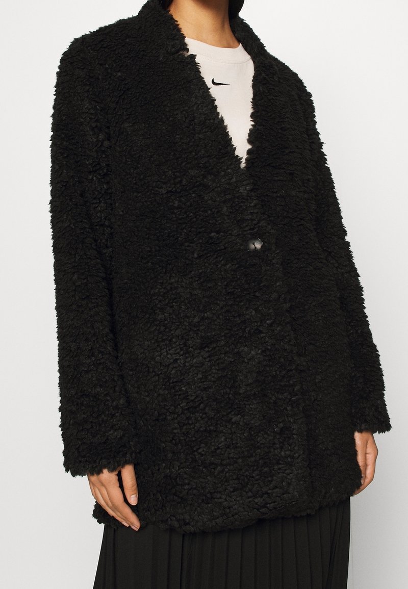 Manteau en fausse fourrure noire avec une surface texturée, de larges revers et une fermeture à un bouton, porté par-dessus une chemise claire et une jupe plissée.