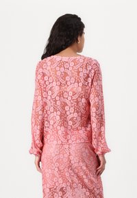 Rosa spetsblus med långa ärmar, blommönster och draperade manschetter; semitransparent tyg med rak skärning och rund halsringning.