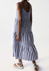 Pruhované bezrukávové šaty v modré a bílé barvě s výstřihem do V, nařaseným živůtkem a volánkovým lemem. V kombinaci s bílými sandály na tenkých řemíncích.