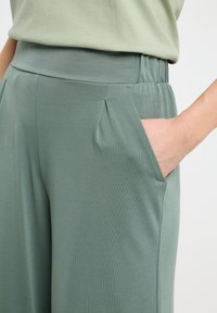 Un pantalon vert clair avec une texture lisse présente une ceinture large, des poches latérales et des détails de plis pour un ajustement décontracté.