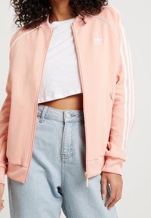 Frau trägt eine hellrosa Adidas Trainingsjacke über einem weißen Crop-T-Shirt und hellblauen hochgeschnittenen Jeans vor einem einfarbigen Hintergrund.
