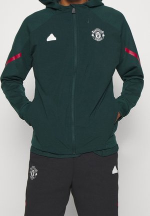 Personne portant un sweat à capuche zippé vert foncé et un pantalon noir avec les logos de Manchester United et Adidas, mains dans les poches, sur fond clair.