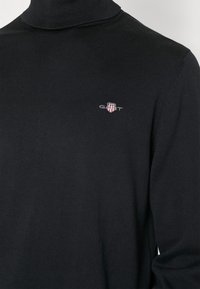 Svart polotröja i ribbad tyg. Har en liten broderad logotyp på vänster bröst i röd, vit och blå.