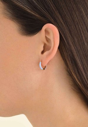 Orecchini a cerchio in tonalità oro con un accento bianco lucido, che mostrano una forma curva. Ambientati su un tono di pelle neutro e capelli lisci.