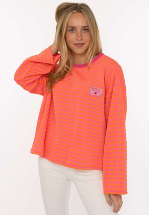 Frau mit langen blonden Haaren, die ein helles orange-pink gestreiftes Langarmshirt und weiße Hose trägt, posiert mit der Hand in der Nähe ihres Kopfes.