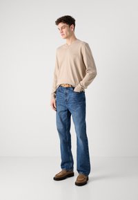 Bezs színű, hosszú ujjú V-kivágású póló, kék egyenes szabású farmer, barna slip-on cipők. A modell áll, kezét a farmer zsebébe dugva, egy sima háttér előtt.