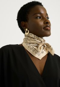 Pañuelo de seda beige con patrones de estrellas y sol en negro, atado alrededor del cuello, combinado con pendientes de aro dorados, usado sobre una blusa negra.