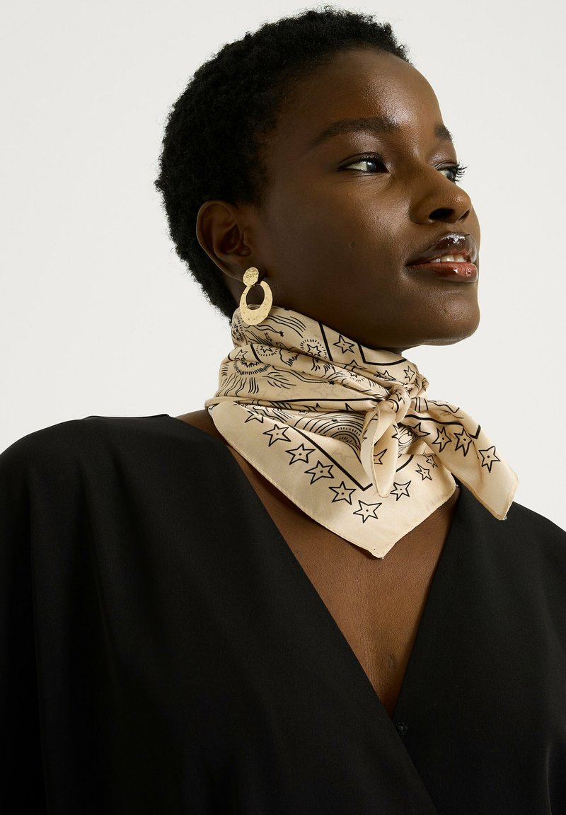 Pañuelo de seda beige con patrones de estrellas y sol en negro, atado alrededor del cuello, combinado con pendientes de aro dorados, usado sobre una blusa negra.