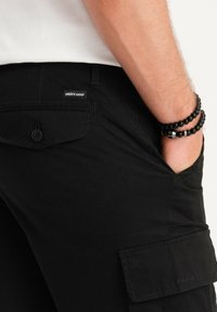 Pantaloni cargo neri realizzati in tessuto resistente, con una tasca a patta dotata di chiusura a bottone, e un'etichetta con la scritta "ombre wear."