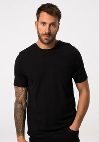 Černé tričko s krátkým rukávem vyrobené z hladkého materiálu, se jako kapsou na hrudi a kulatým výstřihem. Model ukazuje neformální postoj.