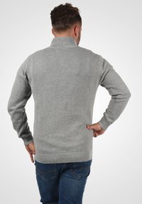 Grauer gestrickter Pullover mit hohem Kragen und gerippten Bündchen, der eine schlichte Textur aufweist. Getragen mit blauen Jeans. Rückansicht.