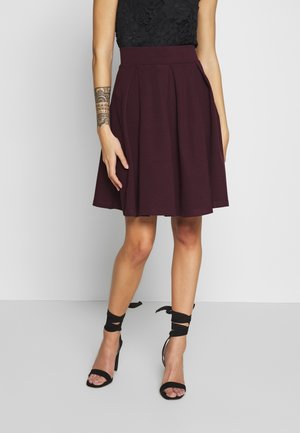 Jupe plissée bordeaux avec une taille haute, associée à des talons à lanières noirs. La jupe a une texture lisse et tombe au-dessus du genou.