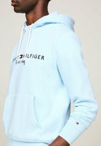 Hombre con sudadera ligera azul de Tommy Hilfiger con bolsillo frontal y texto del logo, vista lateral parcial sobre fondo neutro.