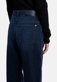 Jeans in denim blu scuro con un design a gamba dritta, tasche posteriori e una toppa in pelle sulla vita. Texture liscia con dettagli hardware minimi.