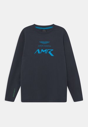Chemise bleu marine à manches longues avec logo Aston Martin AMR bleu sur le devant et petit texte vert « Hackett » sur le poignet de la manche gauche.