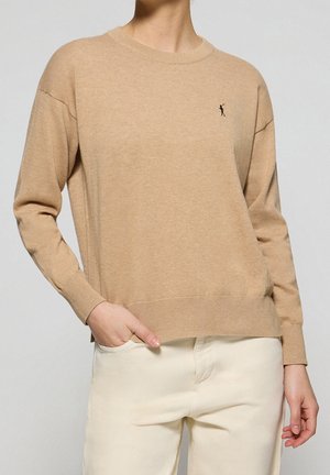 Persona che indossa un maglione beige a maniche lunghe con una piccola figura nera ricamata sul petto e pantaloni color crema chiaro, con la mano in tasca.