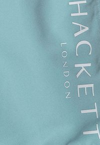 Tessuto blu acquamarina con testo in rilievo bianco "HACKETT LONDON." Il materiale ha una texture liscia senza motivi o accenti visibili.