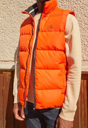 Homme portant un gilet sans manches matelassé orange vif sur une chemise beige à manches longues, debout contre un mur texturé.