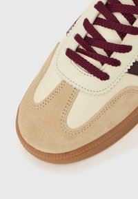 Bout de basket en daim beige et crème avec des lacets grenat et une semelle en caoutchouc marron, couture détaillée, sur fond blanc.