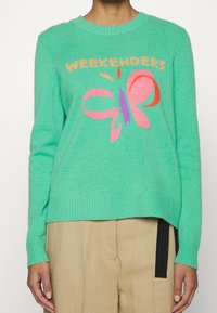 Groene gebreide trui met een geribbelde zoom, met de tekst "WEEKENDERS" en een kleurrijke vlinderafbeelding in roze, oranje en paars.