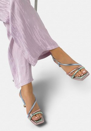 Pies con sandalias de tacón abierto con tiras en tonos metálicos pastel y dedos cuadrados, vistiendo pantalones anchos texturizados en color púrpura claro sobre un fondo blanco.