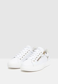Zapatillas blancas con un panel lateral tejido texturizado y acentos dorados. Presentan un cierre de cordones en la parte frontal y un detalle decorativo de cremallera. Suela de goma.