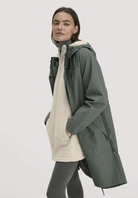 Grüne wasserdichte Jacke mit Kapuze, über einem beigen Fleece-Oberteil getragen. Verfügt über Seitentaschen und ein langes, lockeres Design.