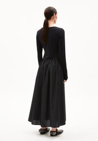 Schwarzes langärmliges Kleid mit figurbetontem Oberteil und gerafftem Rock. Der glatte Oberstoff geht in einen strukturierten, fließenden unteren Teil über und wird mit schwarzen Schuhen kombiniert.