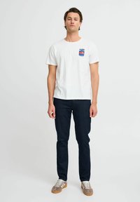 Vit bomull T-shirt med en liten blå och röd grafik på bröstet, kombinerad med mörkblå slim-fit jeans och beige sneakers.