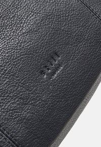 Superficie in pelle nera testurizzata con una grana sottile, caratterizzata da marchi del brand in rilievo e bordi cuciti puliti.