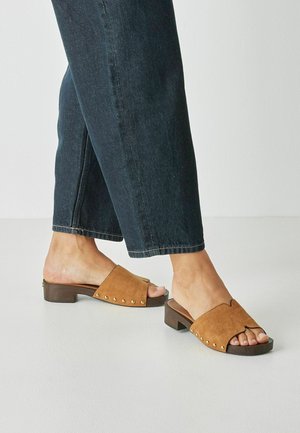 Sandales à talons blocs en daim marron avec un bout ouvert, présentant un design festonné et des clous dorés sur le côté, portées avec un jean denim foncé.