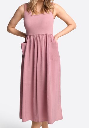 Frau trägt ein ärmelloses, pinkes Midikleid mit geripptem Oberteil, geraffter Taille und großen Fronttaschen, steht mit den Händen in den Taschen.