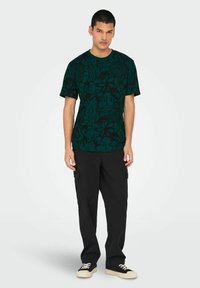Camiseta de color teal con un patrón floral negro, de manga corta y cuello redondo, combinada con pantalones cargo negros y zapatillas.